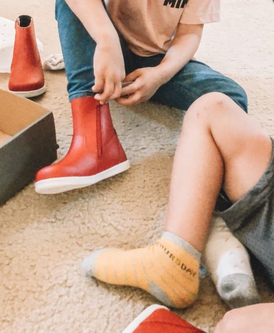 Girl putting on Bobux Paddington Red Boots in bedroom