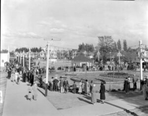 black and white photo of people . Spectators in the front are viewing the action on a circular track. While in the back is the tall steel structure spinning the Swing ride. Tall light standards each support two distinctive lights and bulb lights are strung between the standards for extra evening illumination.