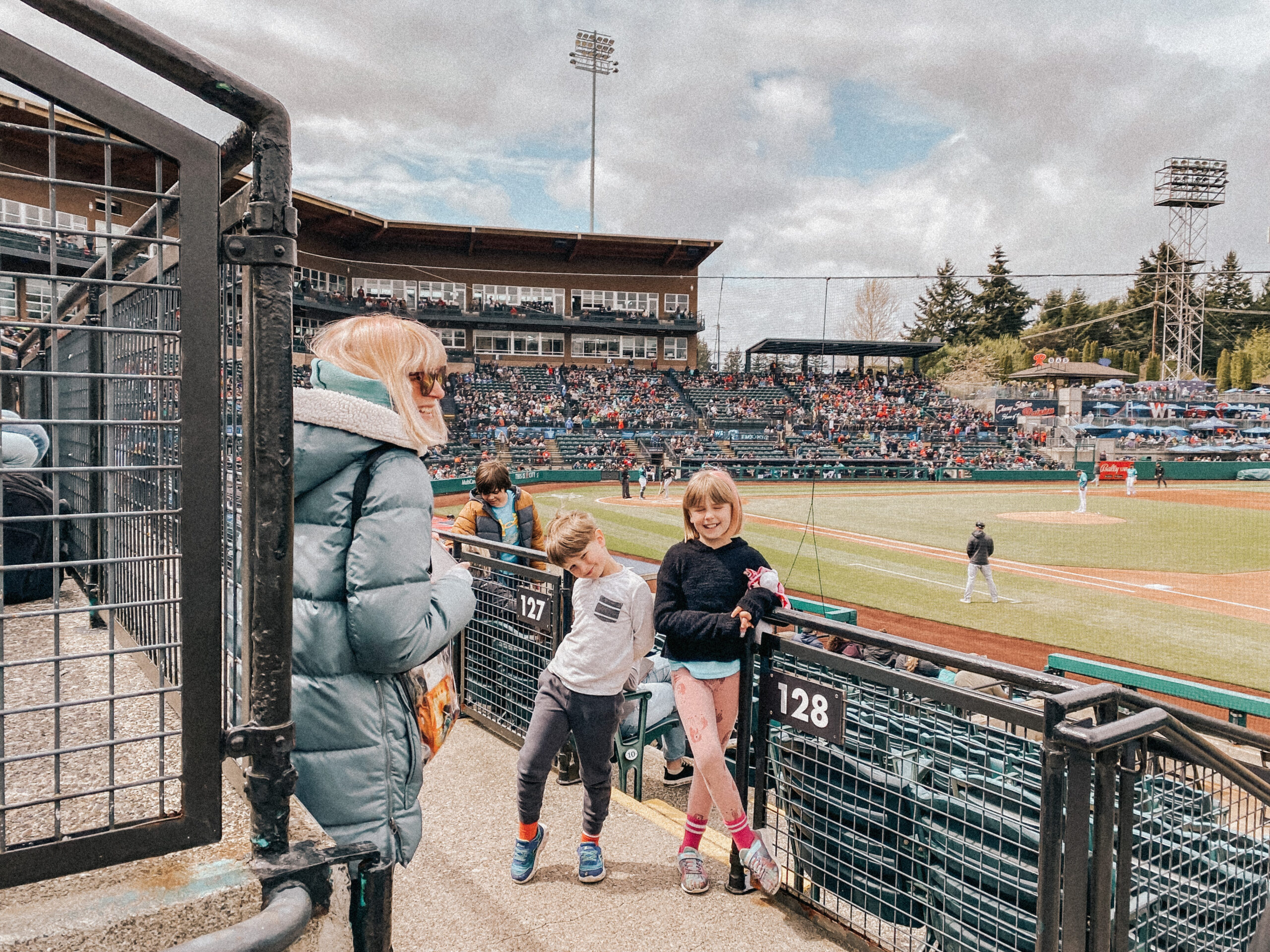 mom in blue coat standing at baseball game with kids